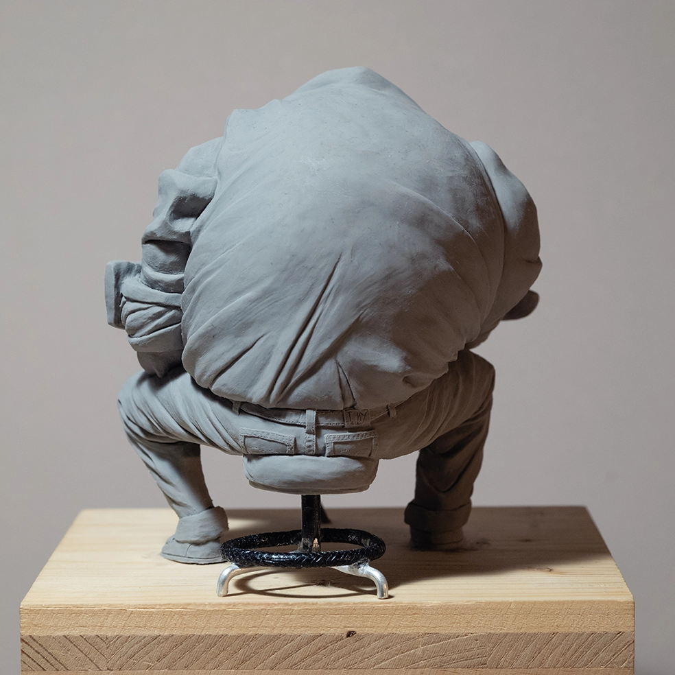 Sculpture by artist Teddy Ros "Café-Créa" 2022, sculpted in polymer clay, depicting a large character sitting on a stool writing in a small notebook, while his coffee cup shatters. Teddy Ros is a contemporary art artist.