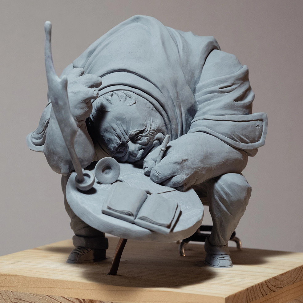 Sculpture by artist Teddy Ros "Café-Créa" 2022, sculpted in polymer clay, depicting a large character sitting on a stool writing in a small notebook, while his coffee cup shatters. Teddy Ros is a contemporary art artist.